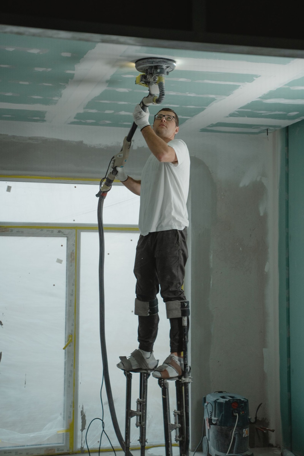 Finisher on stilts sanding a drywall ceiling in a new room